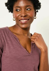 V-neck, short-sleeve top in mauve fabric with a soft texture, featuring a button placket and subtle stitching details at the neckline.