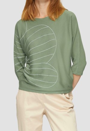 Femme portant un haut à manches longues vert olive avec des lignes abstraites blanches et un pantalon couleur crème, debout avec une main dans la poche.