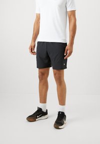 Homem de calções de desporto pretos, meias brancas até ao tornozelo, sapatos de desporto pretos e uma camisa branca de mangas curtas, em pé sobre um chão branco liso.