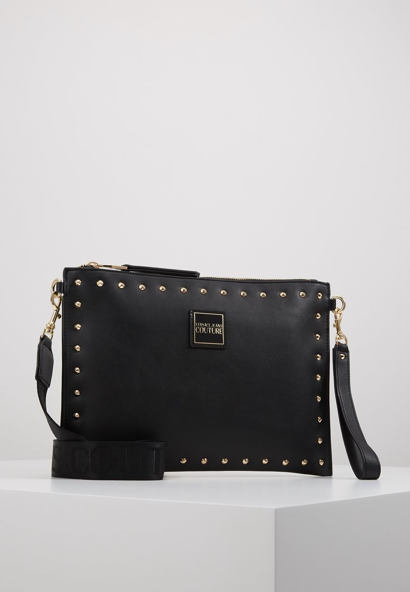 Pochette rectangulaire noire avec bordure en clous dorés, plaque logo Versace Jeans Couture, bracelet poignet amovible et large bandoulière.