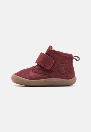 Stivale alla caviglia in suede bordeaux con strap in velcro, interno imbottito e suola in gomma. Presenta un logo circolare sul lato.