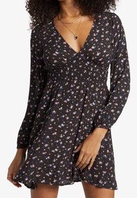 Žena v černých květovaných šatech s dlouhými rukávy, s nařaseným pasem a výstřihem do V, doplněná o vrstvené zlaté náhrdelníky a prsteny.