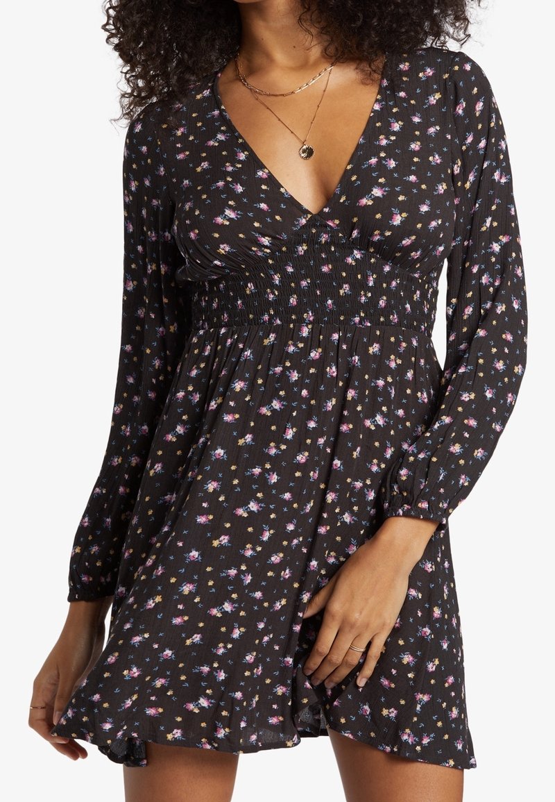 Žena v černých květovaných šatech s dlouhými rukávy, s nařaseným pasem a výstřihem do V, doplněná o vrstvené zlaté náhrdelníky a prsteny.