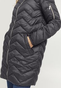 Sort polstret jakke med chevron-quilting, frontlynlås, sidelommer og et slankt design. Stoffet fremstår glat med en let glans.