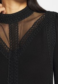 Svart topp med genomskinlig meshsektion och intrikat svart spetskant längs halsringningen och framsidan, vilket skapar en lagerdesign-effekt.