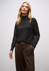 Schwarzer Rollkragensweater aus einem weichen Material, kombiniert mit braun gemusterten Hosen, die über einen Knopfdetail und seitliche Taschen verfügen.