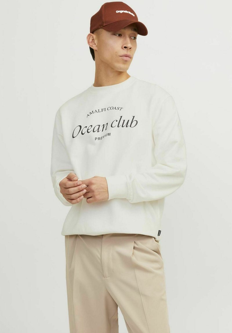 Felpa bianca con testo nero "Ocean Club" e dettagli grafici aggiuntivi, abbinata a pantaloni beige e un berretto marrone. Materiale misto cotone.