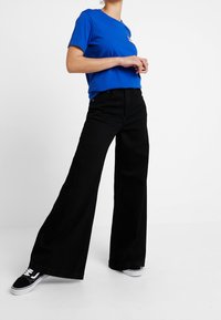 Blå kortärmad t-shirt i kombination med hög midja svarta vida byxor och svarta sneakers. Outfiten har en avslappnad passform och slät textur.