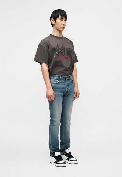 Γκρι γραφικό μπλουζάκι με μαύρο και κόκκινο κείμενο, συνδυασμένο με ξεθωριασμένα μπλε τζιν και ασπρόμαυρα ψηλά αθλητικά παπούτσια.