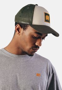Gorra trucker verde y beige con un logo de elefante cuadrado; parte trasera de malla transpirable; combinada con una camiseta de cuello redondo gris que presenta un pequeño elefante.