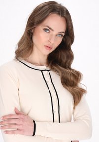 Cremfarbener Cardigan mit schwarzem Besatz, Perlenknopfdetails, langen Ärmeln und weicher Textur. Entworfen für eine figurbetonte Silhouette.