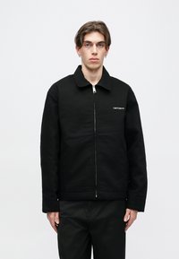 Μαύρο μπουφάν με φερμουάρ και κολάρο, κατασκευασμένο από υφασμάτινο υλικό με υφή, με μια μικρή λευκή λογότυπο στο αριστερό στήθος και ίσια γραμμή.