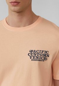 Peachkleurig katoenen t-shirt met een ronde hals en een marineblauw grafisch ontwerp met de tekst "PACIFIC CUSTOMS VENICE BOARDS&KITS" op de borst.