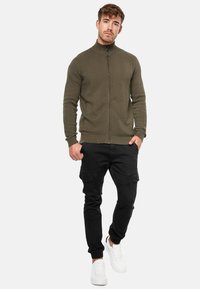 Oliven grøn zip-up jakke med et struktureret chevronmønster, parret med sorte cargo bukser og hvide sneakers. Casual pasform.