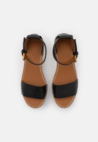 Sandales en cuir noir avec de larges brides, semelle intérieure beige et semelle en tressé. Une bride de cheville ajustable avec des détails en métal doré.