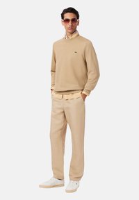 Sudadera beige con cuello redondo, combinada con una camisa amarilla clara con cuello y pantalones beige, acompañada de zapatos blancos. Gafas de sol marrones.