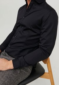 Svart långärmad skjorta med skjortkrage, tillverkad av mjukt tyg, med en figursydd design och subtila knappdetaljer. Bärs med strukturerade grå byxor.