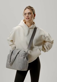 Borsa a spalla grigia imbottita con una finitura testurizzata, dotata di una tracolla regolabile e dettaglio arricciato sul bordo superiore, abbinata a un hoodie color crema.