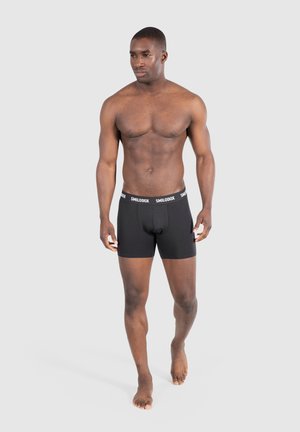 Bañadores negros con una cinturilla blanca "SMILODOX", hechos de una tela suave, que se ajustan cómodamente, usados por un modelo masculino de pie descalzo.