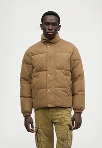 Bruine corduroy pufferjack met een hoge kraag, voorste drukknoopsluiting en twee zijzakken. Gestoeld met groene cargobroek.