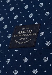 Tummansininen kangas valkoisilla pilkuilla, jossa on musta etiketti valkoisella tekstillä, jossa lukee "GAASTRA SAILMAKER SUPPLIES SNEEK ALANKOMAAT."