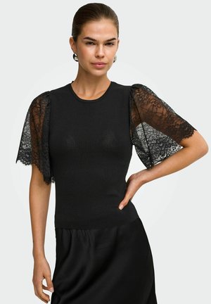 Femme portant un haut noir cintré avec des manches courtes en dentelle transparente et une jupe noire, debout avec une main sur la hanche devant un fond uni.