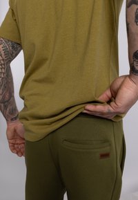 Olivenfarbener T-Shirt aus weichem Stoff, kombiniert mit passenden olivenfarbenen Hosen mit einer Seitentasche und einem Markenetikett.
