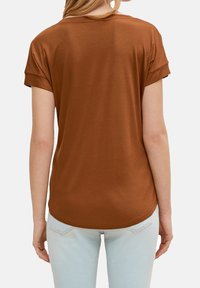 Braun kurzärmliges Oberteil mit umgeschlagenen Ärmeln, weicher Textur, abgerundetem Saum und lockerer Passform. Getragen mit hellblauen Jeans.