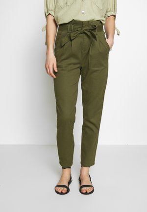 Femme portant un pantalon taille haute vert olive style paperbag avec une ceinture nouée, associé à des sandales noires à bouts ouverts et une chemise vert clair.