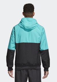 Man in een zwart en turquoise hoodie-jack, staand met zijn rug naar de camera tegen een eenvoudige lichte achtergrond.