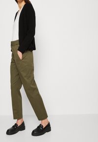 Zwarte gebreide cardigan, witte v-hals t-shirt, olijfgroene broek met zijzakken en slanke zwarte loafers met een dikke zool.