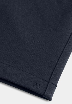 Tissu tricoté gris foncé avec couture visible et petit logo circulaire brodé près de l'ourlet sur un vêtement.