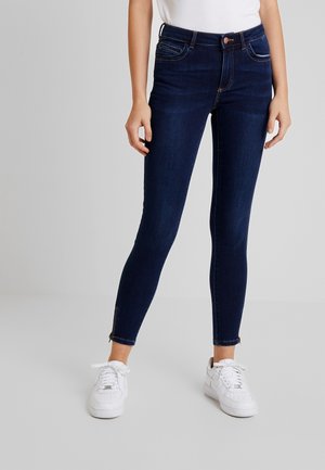 Osoba ubrana w ciemnoniebieskie dopasowane dżinsy z zamkami przy kostkach, białe trampki i biały top, stojąca na jednolitym jasnoszarym tle.