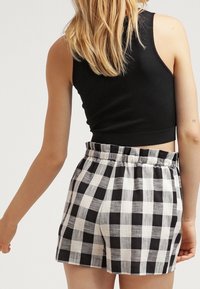 Nainen, joka on pukeutunut mustaan ilman hihoja cropped topiin ja korkeavyötäröisiin musta-valkoisiin ruudullisiin shortseihin, näkyy takaa vaaleat hiukset kammattuna.