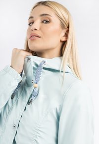 Giacca impermeabile azzurro chiaro con colletto alto, cordini regolabili a righe e dettagli argentati. Tessuto liscio e logo sottile.