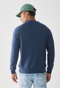 Man met een groene pet, marineblauwe sweatshirt en lichtblauwe spijkerbroek, met zijn rug naar de camera gekeerd tegen een effen lichte achtergrond.