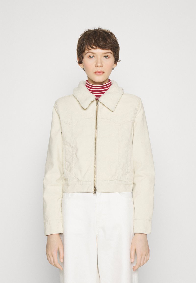Veste beige coupée avec un col en peluche, fermeture éclair sur le devant et texture matelassée, associée à un col roulé rayé rouge et blanc en dessous.