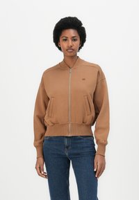 Chaqueta bomber marrón con cierre de cremallera frontal, puños y dobladillo acanalados, y bolsillos laterales. Tejido suave con un acabado ligeramente texturizado.
