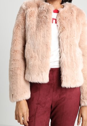 Personne portant une veste en fausse fourrure rose clair sur un t-shirt blanc avec un texte rouge, associée à un pantalon taille haute rouge foncé.