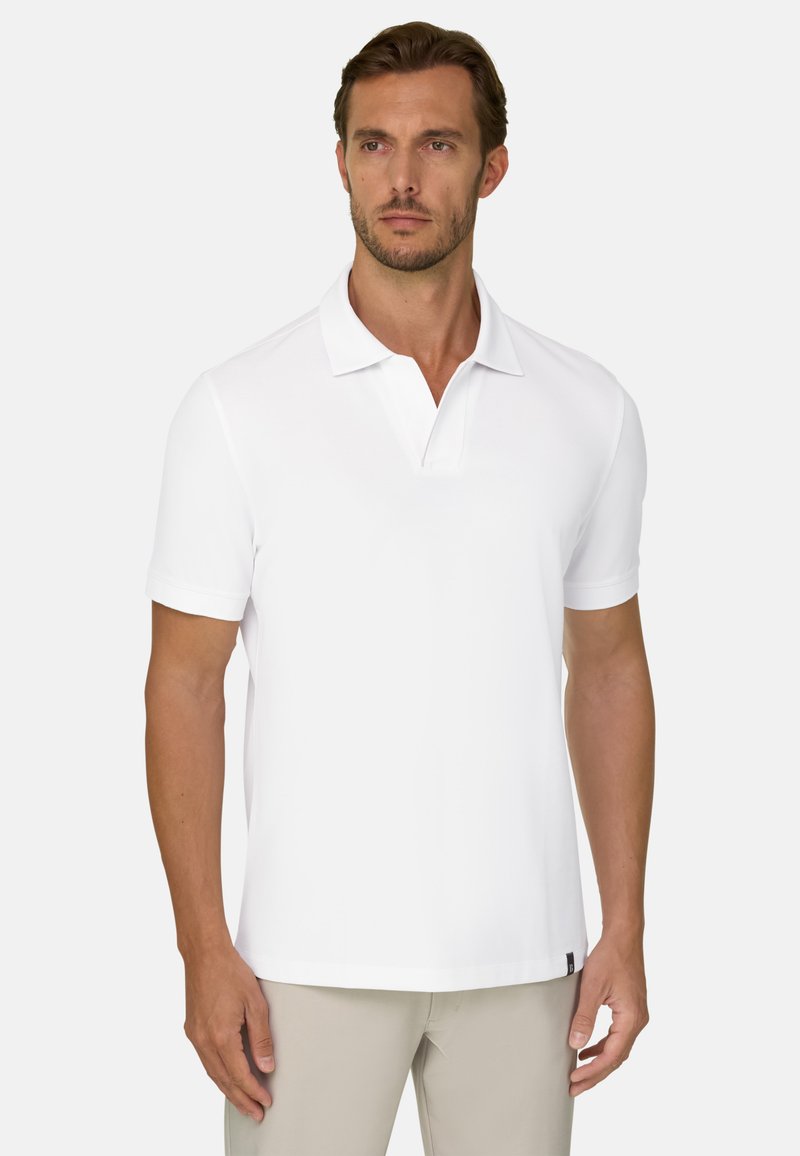 Homme aux cheveux bruns courts et à la barbe claire, portant un polo blanc uni à manches courtes et un pantalon beige sur un fond clair.