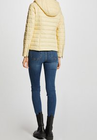 Light yellow puffer jacket with a hood, detailed quilting, and zippered sides, paired with dark blue skinny jeans and black ankle boots.