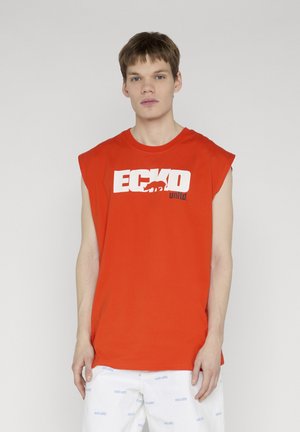 Mladić s kratkom kosom nosi crvenu majicu bez rukava s velikim bijelim logotipom "ECKO" i bijele kratke hlače s uzorkom svijetloplavog teksta.