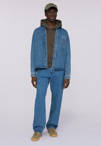 Jeansjacka, blå, med bröstficka; olivgrön hoodie under; blå jeans; beige skor; accessoriserad med en ljusgrön keps.