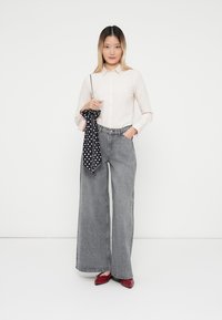 Jasnoróżowa koszula zapinana na guziki, szare szerokie dżinsy, czerwone buty i czarna chusta w kropki. Model stoi na tle białym.