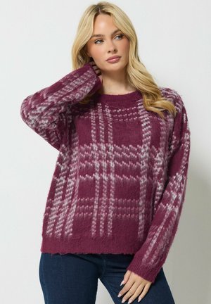 Femme aux cheveux blonds portant un pull à carreaux violet duveteux et un jean bleu foncé, posant avec une main près du cou.