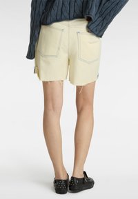 Shorts en denim jaune avec un ourlet effiloché, comportant deux poches arrière soulignées de couture bleue. Associés à des chaussures à enfiler noires à clous.