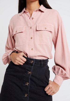 Lys pink skjorte med knapper og to forlommer, parret med en mørk denim blyantskirt med frontknapper. Skjorten har lange, bløde ærmer.