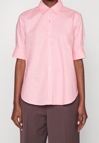 Kortärmad, knäppt skjorta i ljusrosa bomull. Har en spetsig krage och en diskret logotyp på bröstet. Rakt snitt med rundad nederkant.