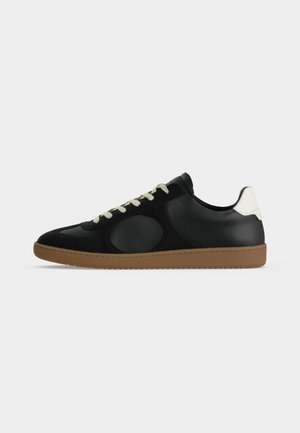 Zwarte lage sneaker met wit hieldetail, beige veters en bruine rubberen zool, zijaanzicht op een witte achtergrond.