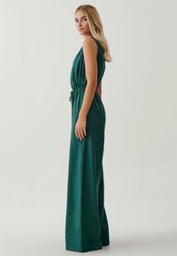 Tuta verde senza maniche con vita aderente, gambali larghi e una trama liscia. Presenta un cordino in vita per la regolazione.